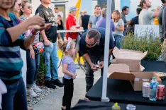 A street event with people in the background, a little girl interacting with a man by a table with pizza boxes, while another person captures the moment on their phone.