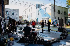 A live band performs on a street stage in front of an audience during a sunny outdoor event, with buildings and urban street art in the background.
