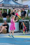 A young girl in a pink dress playing with a colorful ribbon toy at a street fair with people in the background near a tent labeled "HOGMOLLYBREWING.COM"