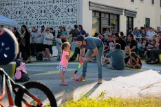 A man crouching down to show a colorful toy to a young girl at a lively outdoor community event with onlookers in the background.