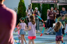 Children playing with colorful ribbons at a street fair with onlookers in the background.