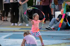 Young girl playing with hula hoop outdoors at an event while another child is sitting on the ground nearby.