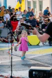 A small child in a pink dress dancing on a street with a microphone stand in front and an audience in the background.