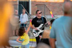 Musician playing a white electric bass guitar interacts with a young child in a yellow shirt at an outdoor event with onlookers in the background.