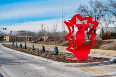 Red abstract sculpture at a road roundabout with bare trees and a blue sky in the background.