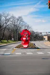 Alt: A red sculpture with circular elements located in the center of a roundabout under a clear blue sky.