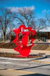 Red abstract sculpture in an outdoor setting with bare trees and a clear blue sky in the background.