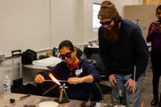 Woman wearing safety glasses using a torch to heat glass while smiling man with a beard watches and another person observes with a smartphone in the background.