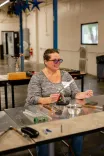 Woman smiling while working with glass rods at a workbench in a workshop setting.