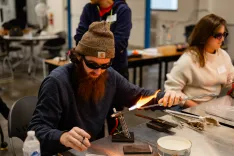 Man with a beard wearing a beanie and safety glasses shaping glass with a blowtorch in a workshop setting.