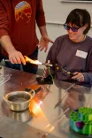 A smiling woman wearing safety glasses and using a blowtorch on a piece of metal in a glassworking workshop, assisted by a person in a red shirt.