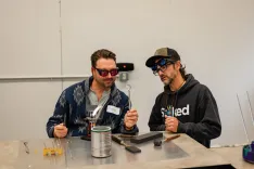 Two people wearing safety glasses working with glassblowing tools at a workshop table.