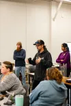 Instructor speaking to students in a classroom setting, with attentive participants looking on.