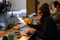 Person wearing safety glasses and a beanie doing glassblowing with a flame torch in a workshop environment.