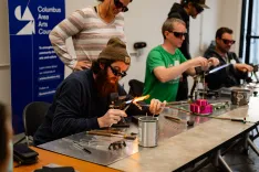 Group of people wearing safety glasses working with flame torches during a glass blowing workshop.
