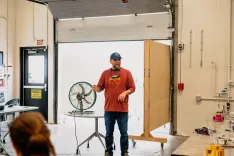 Man in a baseball cap and glasses speaking during a workshop in a garage-like room with an open garage door, tables with tools, and a metal fan.