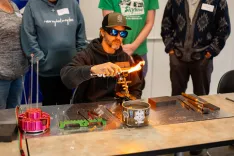 Glassblower demonstrating techniques to an attentive audience, heating a glass rod over a gas burner, surrounded by glassblowing tools and safety equipment.
