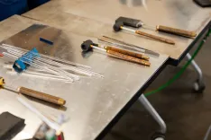 Various glass blowing tools laid out on a metallic worktable, including clear glass rods, wooden handles with metal tips, and graphite paddles.