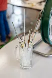 A clear glass mug filled with various paintbrushes on a white surface with blurred background.