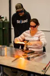 A woman with protective glasses working on a metal piece using a blowtorch with a man supervising in the background.