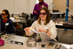 Woman in safety goggles laughing while working with a gas torch on a glass blowing activity in a workshop with another person in the background.