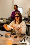 Woman in a cream sweater and sunglasses laughing while shaping a glass piece with a torch as another woman looks on in a workshop environment.