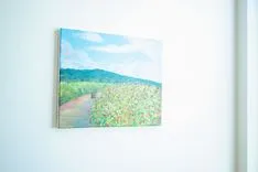 Landscape painting hanging on a white wall, depicting a rural scene with a path through a green field leading towards distant hills under a blue sky.