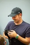 Man in a cap smiling and holding a small black kitten with a blurred background.