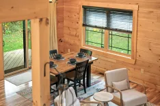 Cozy wooden cabin interior with a dining area set for four, modern furniture, and a view of the outdoors through windows.
