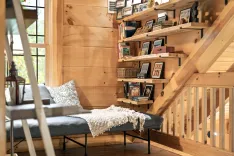 Cozy wooden cabin interior with a staircase lined with bookshelves full of books and framed photos, and a window seat with cushions and a throw blanket.