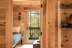 Cozy wooden cabin interior with a view of the forest through the glass door, featuring a bed and wall-mounted lantern.