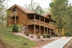 A rustic wooden cabin with a covered front porch and an American flag hanging on the wall, surrounded by trees with autumn leaves and a landscaped garden.