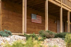 An American flag hanging on the wooden wall of a covered porch with landscaping including shrubs and small rocks in the foreground.