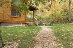 A cozy wooden cabin with a porch in an autumnal forest setting, featuring a fire pit and benches on a leaf-covered lawn.