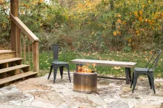 Outdoor patio area with a fire pit, two chairs, and a bench amidst autumn foliage.