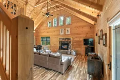 Interior of a cozy cabin living room with wooden walls and ceiling, plush sofa, stone fireplace, and large windows.