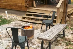 Outdoor patio scene with a lit fire pit, two metal chairs, and a wooden bench near a log cabin.