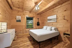 Cozy cabin bedroom interior with wooden walls and ceiling, queen-sized bed, framed artwork, wall-mounted guitar, and window with nature view.