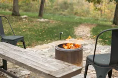 A cozy outdoor fire pit with flames surrounded by chairs and a wooden picnic table in an autumnal setting.