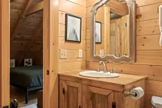 Rustic wooden bathroom interior with a framed mirror above a wooden vanity, a glimpse of a bedroom area in the background, and modern amenities.