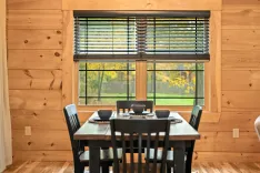 A cozy dining area with a wooden table set for four in front of a large window with blinds, overlooking autumn trees.