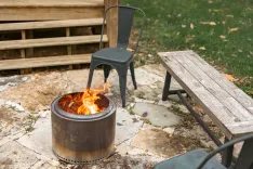 Outdoor setting with a metal fire pit ablaze, a single metal chair, and a wooden bench on a stone-paved ground with grass and leaves in the background.