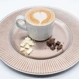 A cup of cappuccino with heart-shaped latte art on a ribbed bronze plate with pieces of white and dark chocolate.