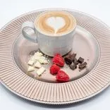 Cup of cappuccino with heart-shaped latte art on a textured plate accompanied by pieces of white and dark chocolate, and fresh raspberries.