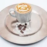 A cup of caramel macchiato with whipped cream and caramel topping on a beige plate garnished with shredded coconut and chocolate pieces.