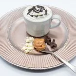 A hot beverage topped with whipped cream and chocolate syrup in a cup on a saucer, white chocolate pieces, dark chocolate pieces, and a dollop of peanut butter on a spoon, all on a textured pink plate.