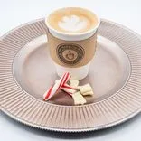 Paper coffee cup with latte art on a textured ceramic plate accompanied by a candy cane and pieces of white chocolate.