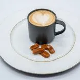 Black cup of cappuccino with heart-shaped latte art on a white plate with caramel pieces, on a white background.