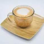 A cup of cappuccino with frothy milk on top, on a bamboo plate against a white background.