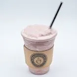 A frothy pink smoothie in a clear plastic cup with a cardboard sleeve and a metal straw, set against a white background.